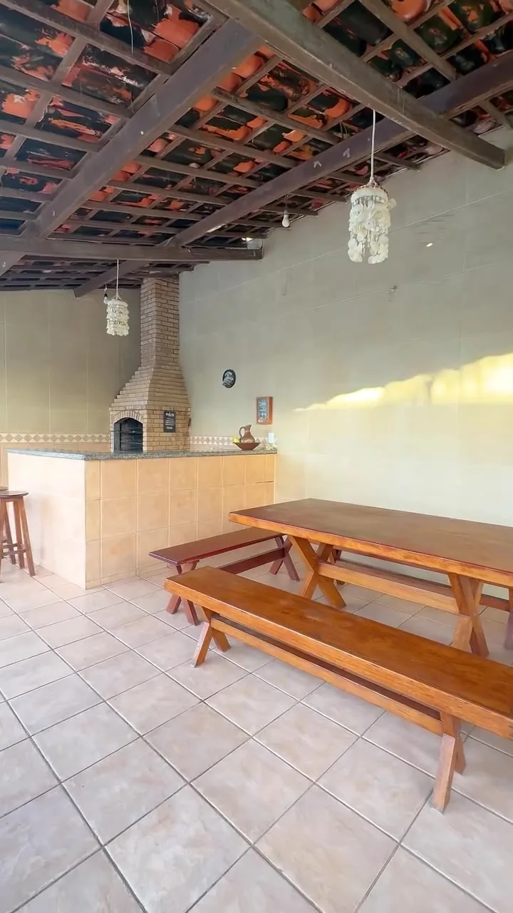 Deck coberto com churrasqueira de alvenaria, mesa comprida de madeira e luminárias de fibra
